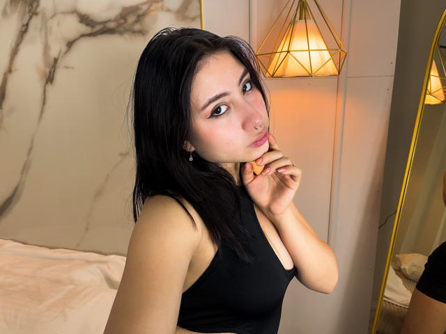 VictoriaAngels with black hair, wearing a black tank top, touching her nose with her right hand, looking at the camera, her nose is pierced, medium tits, brown eyes.