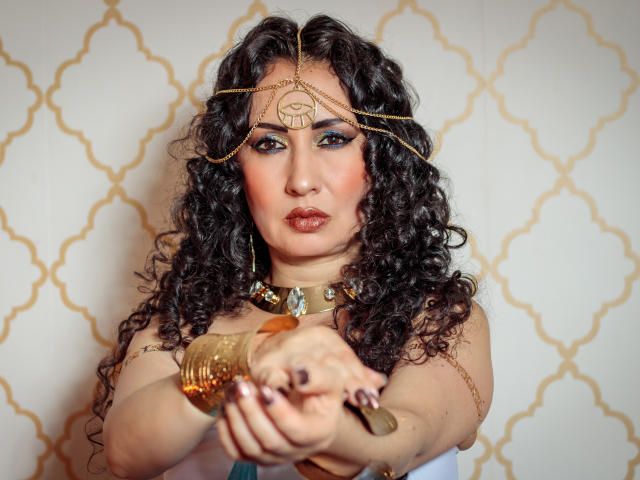 SamanthaAntolin with curly black hair, wearing gold chain headpiece, choker, and bangles, pointing forward, serious expression, blue eye makeup, and brown lipstick. Boobs visible, white top.