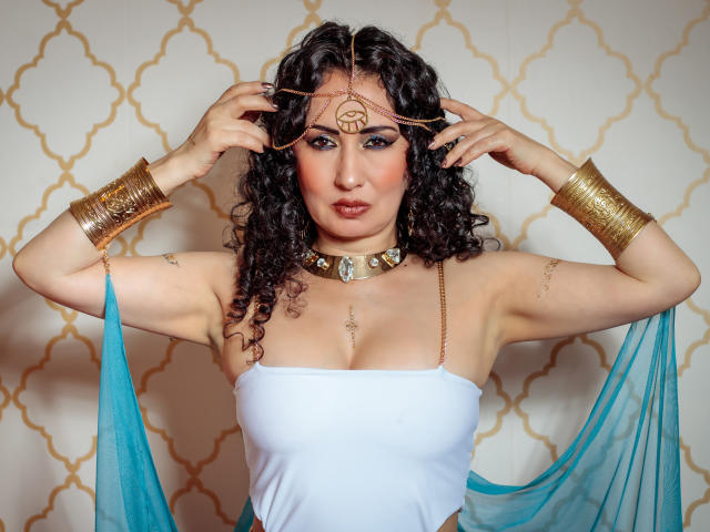 SamanthaAntolin with curly dark hair, wearing a white strapless top, gold headpiece, bracelets, and a turquoise sheer fabric, showing off her tits and a jeweled necklace.