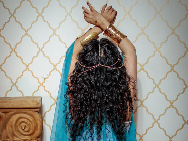 SamanthaAntolin with curly black hair, gold arm bands, and a blue sheer top, arms crossed, back to camera, wearing a gold headpiece, against a patterned wall.