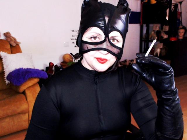 ImperatrizaSanda in black latex cat mask, bodysuit, and gloves, holding a cigarette, smiling with red lipstick, showing off her tits and ass, in a living room.