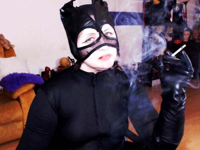 ImperatrizaSanda in a black cat mask and leather outfit, smoking a cigarette, with blue smoke, wearing black gloves, in a cozy room with a brown sofa.