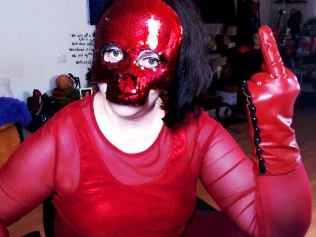 ImperatrizaSanda wearing a shiny red mask with a hole for the mouth, red sheer top, and red latex gloves, giving a middle finger with a raised eyebrow.