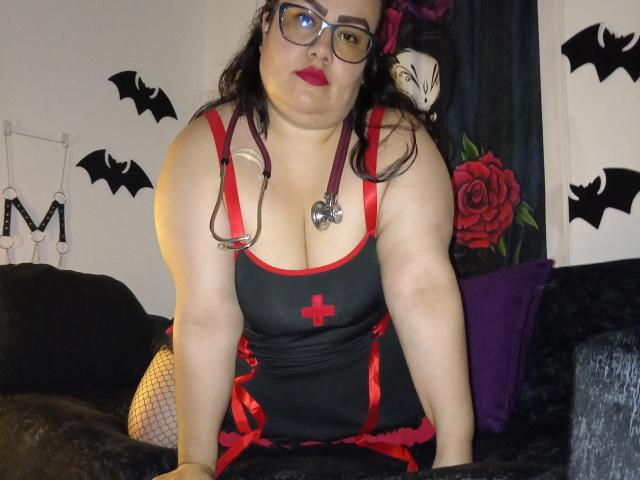 FoxyBell, avec ses joues rondes et son rouge à lèvres rouge, portant des lunettes, en tenue d'infirmière noire et rouge avec une croix, un stéthoscope et des bas résille, penchée en avant sur un canapé noir..