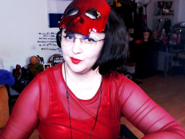 ImperatrizaSanda wearing a red, sheer top and a red, skull mask with black eye holes, has red lipstick and short black hair, with glasses and a black necklace.