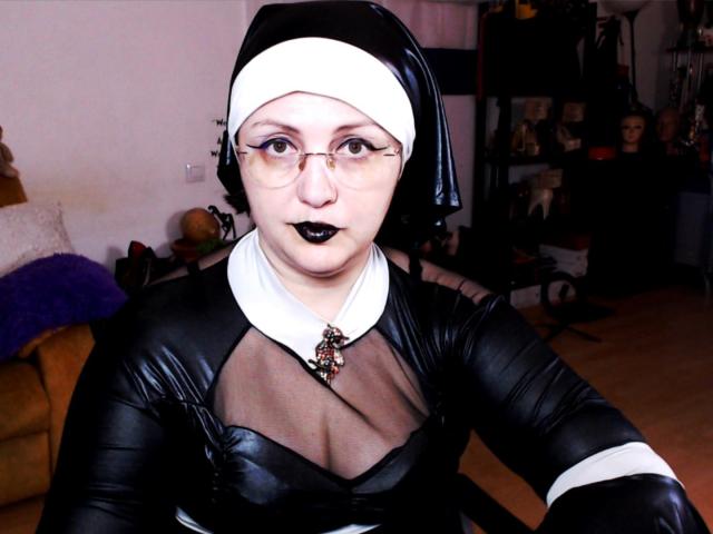 ImperatrizaSanda, wearing a black nun outfit with a white wimple, glasses, black lipstick, and sheer top, showing her tits through mesh. She has a black necklace.