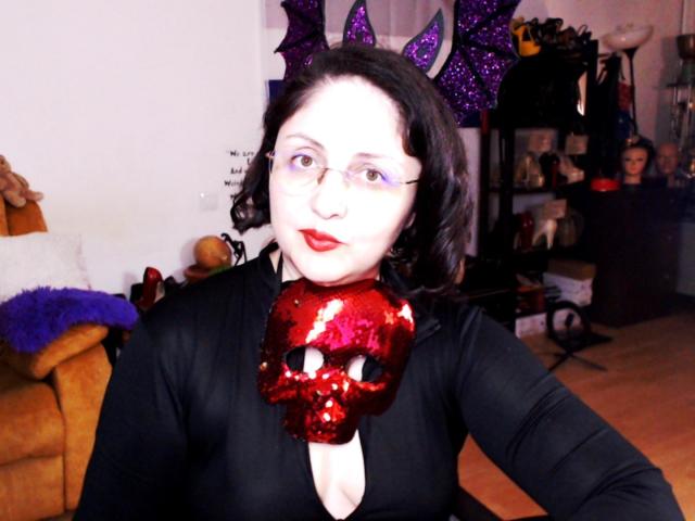 ImperatrizaSanda with pale skin, black curly hair, red sequined skull mask, black top, purple bat wings, red lipstick, glasses, and a cute ass, sitting in a cozy room.