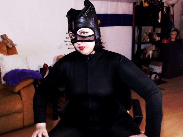 ImperatrizaSanda in a black leather cat mask with ears, black long-sleeve bodysuit, sitting on a chair, wooden floor, red lipstick, black hair, looking at camera.