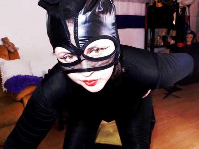 ImperatrizaSanda is a white woman with red lipstick, black latex mask, and cat ears, bending forward, wearing a black latex suit, showcasing her pale face and black-tinted eyes.