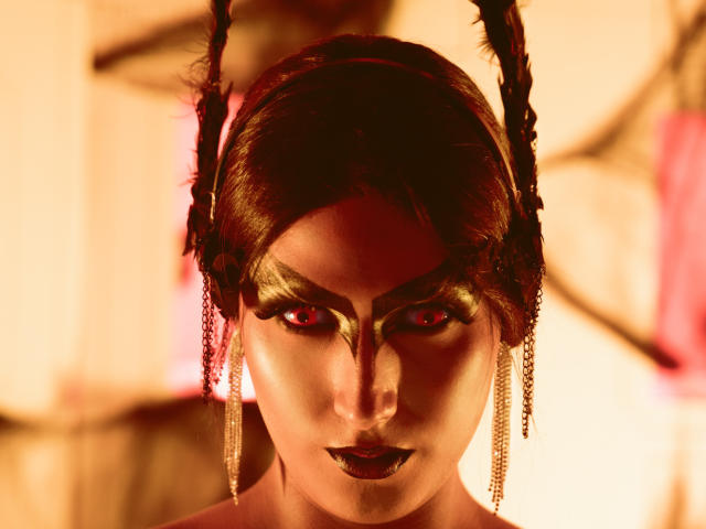 FallonRogers with dark makeup, red eyes, and black lipstick, wearing a headpiece with feathers and chains, looking intense. Her hair is styled in braids, illuminated by warm light.