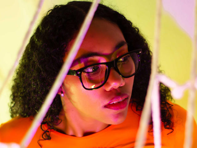 AishaRhodes with curly black hair, wearing black glasses, orange shirt, and pink lipstick, looking through white strings, with purple and yellow lighting on her face.