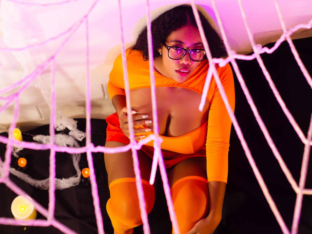 AishaRhodes, with curly black hair and glasses, crouches in front of a pink spider web, wearing an orange, cleavage-revealing, long-sleeve outfit and matching thigh-highs, touching her tits.