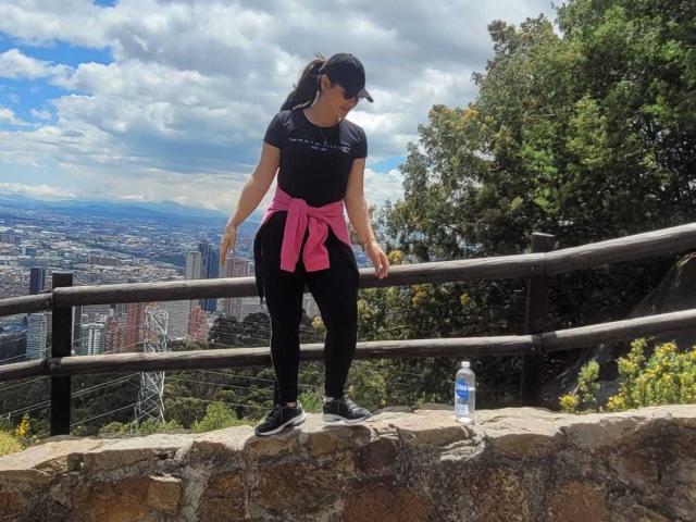 FrancheskaWaalsh stands on a stone ledge, wearing black leggings, black top, pink jacket tied around waist, black sneakers, and black cap. Water bottle on ledge. Tree and railing.