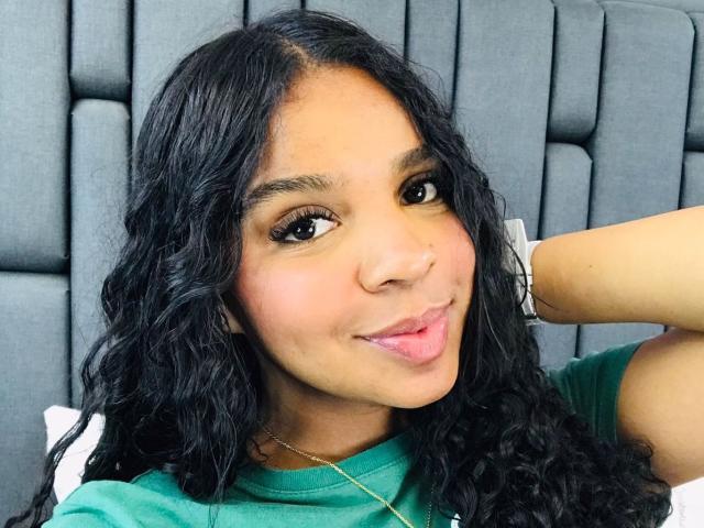 LisaMendez with curly black hair, brown eyes, and green shirt, sticking out pink tongue, sitting against gray padded headboard, arm raised, looking cute.