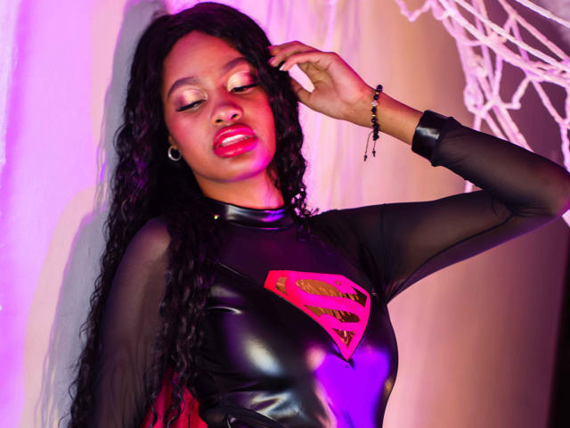 StormiHills with dark curly hair, red lipstick, black sheer long-sleeve top with shiny black latex and pink Superman logo, hand in hair, purple and pink lighting.