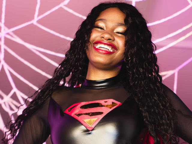 StormiHills with dark curly hair, smiling with red lipstick, wearing a black latex Superman shirt with a red "S" logo, looking happy and cute.