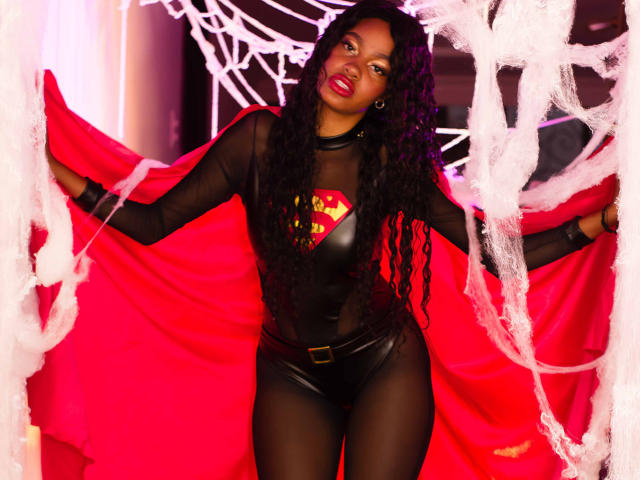StormiHills with long curly black hair, wearing a black mesh Superman costume, red cape, and black leather pants, standing in front of white webbing, smiling with red lipstick.