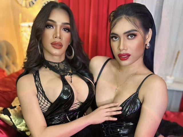 KatherineAmberDuo with big tits in shiny black latex dresses, holding hands, red lips, hoop earrings, long dark hair, and seductive looks, standing in a sexy, intimate setting.
