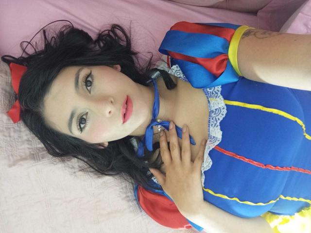JeneKurz lies on a pink bed, wearing a blue Disney-style dress with red and yellow accents, touching her chest with a hand, showing pale skin and black hair.