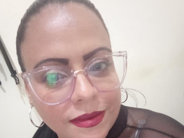BigPussySquirting has clear glasses, red lipstick, and green eye makeup. Her dark hair is pulled back, and she's wearing a black sheer top with silver hoops.