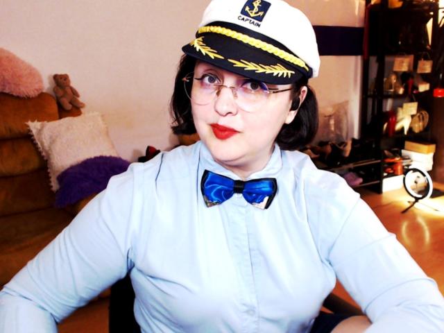 ImperatrizaSanda with glasses, red lipstick, blue shirt, white sailor hat, blue bowtie, and small tits, sitting at a wooden table, looking at the camera.