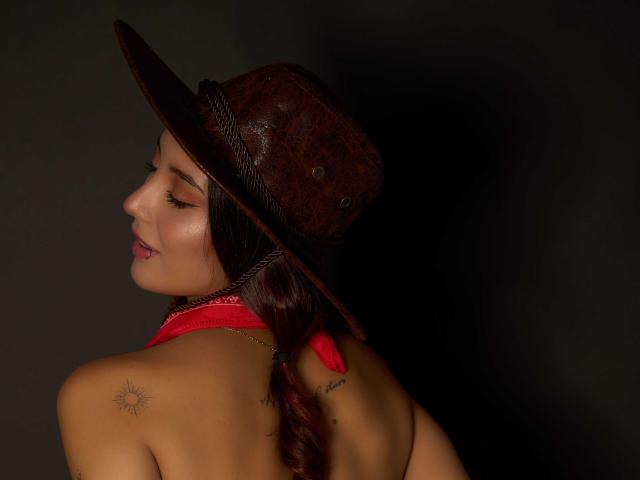 CarlaFranco mit einem braunen Cowboyhut, einem roten Halstuch, geflochtenen roten Haaren, einem kleinen Sonnen-Tattoo auf der linken Schulter, einem leichten Lächeln, im dunklen Hintergrund, mit sichtigem Hintern, die über die Schulter schaut..