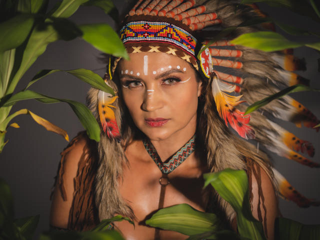 SofiaVerlek with Native American headpiece, white face paint, and feathers, surrounded by green leaves, showing her tits and a serious expression, in a dark, moody photograph.