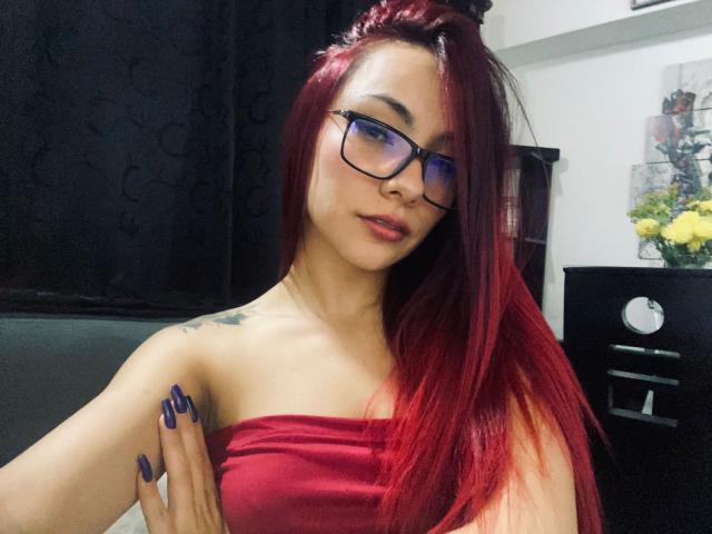 ValeryHarllow with red hair, glasses, and a strapless red top, showing off her tits, tattoo on her shoulder, and blue nails, looking confident.