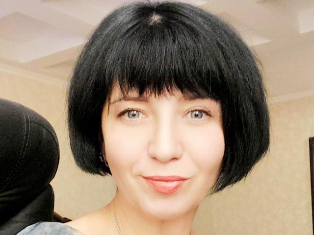 TaniaFire with short black bob, puffy pink lips, and a cute smile, wearing a gray top, has light skin and blue eyes. She's sticking her tongue out slightly.
