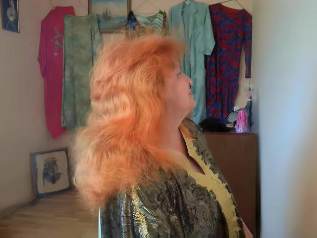 LadaMay, con cabello naranja, ondulado y esponjoso, vestida con una túnica verde metálica brillante, vista de perfil, con un piercing en la nariz, en una habitación con ropa colgada, suelo de madera y un cuadro enmarcado..