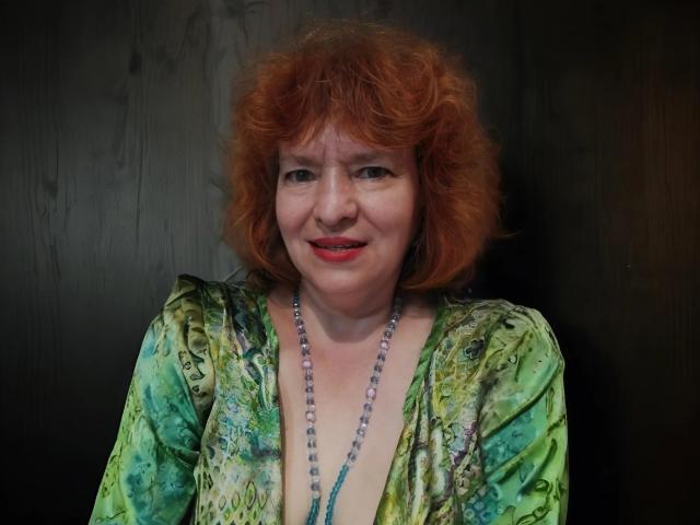 LadaMay heeft krullend rood haar, een jurk met een groen patroon en een kralenketting, en ze glimlacht met rode lippen. Haar borsten zijn licht zichtbaar onder de jurk. Donkere achtergrond..