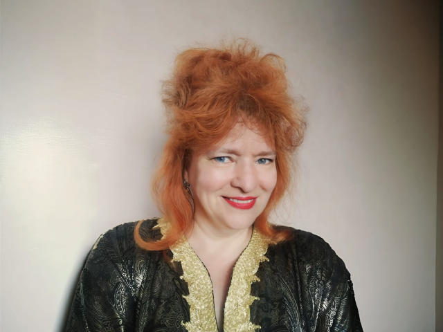 LadaMay with wild red hair, smiling, wearing a black and gold patterned robe, has pale skin and red lipstick, looking slightly to the side.