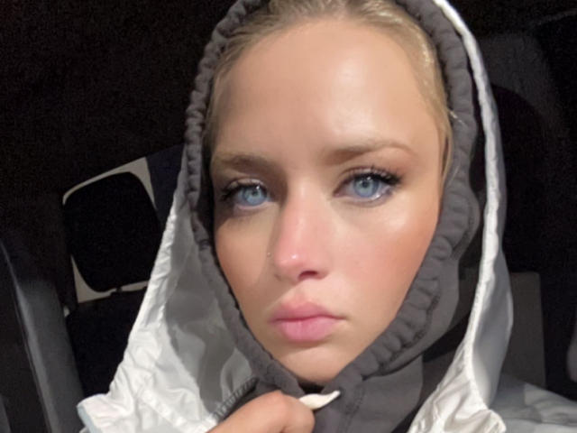 KykyX hat blaue Augen, blondes Haar und ein ernstes Gesicht mit leicht geöffneten, rosafarbenen Lippen. Sie trägt eine weiße Jacke mit Kapuze, und ihre Hand hält den Kragen..