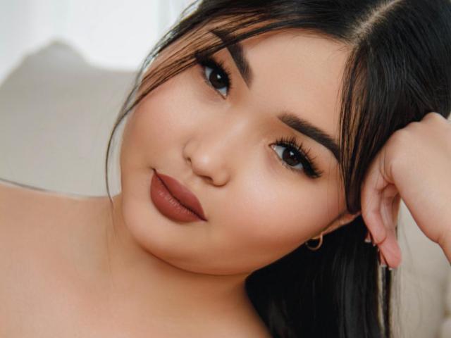 TeyaFaber's face, with dark eyebrows, long eyelashes, brown lipstick, and black hair, has her hand behind her head, showing a cute, slightly sexy expression.
