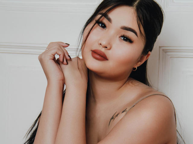 TeyaFaber has long black hair, full eyebrows, dark eyeliner, and red lipstick. She's looking at the camera with a slight smile, arms crossed, showing her tits.