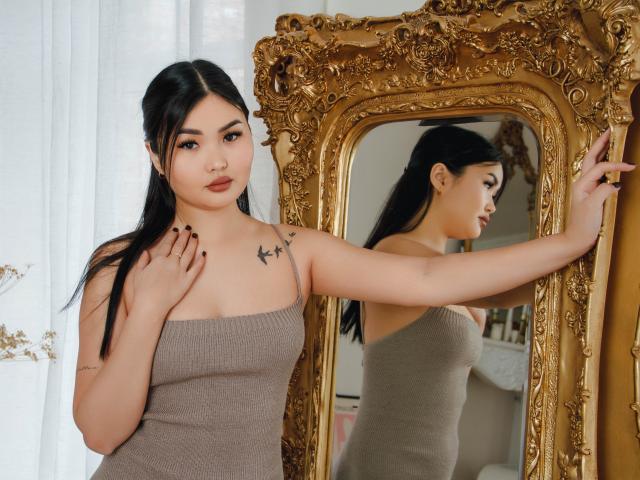 TeyaFaber, met zwart haar, een bruine jurk en kleine borsten, staat voor een sierlijke gouden spiegel, waarbij ze deze met haar rechterhand aanraakt en een tatoeage op haar linker schouder laat zien..