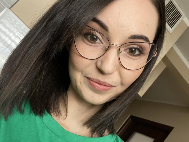 ChloeSensual has pale skin, green eyes, and black shoulder-length hair. She's wearing glasses and a green shirt, with a slight smile and a small acne mark on her chin.