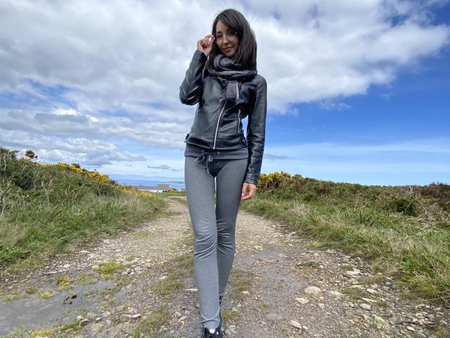 ChloeSensual with dark hair, wearing a black top, gray pants, and a scarf, standing on a rocky path, touching her head, smiling, with a blue sky and clouds.