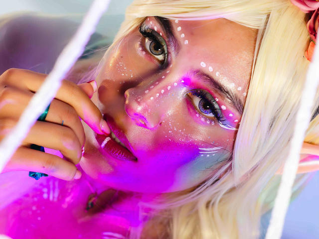 SamanthaEva69 with blonde hair, pink and purple face paint, white dots on forehead, hand near mouth, pink lipstick, pink and purple light, looking into camera, soft expression.