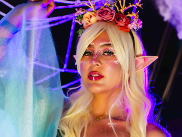 SamanthaEva69 with blonde hair, elf ears, flower crown, glowing face paint, pink lipstick, holding a sheer blue dress, neon purple and blue lighting. Boobs are not visible.