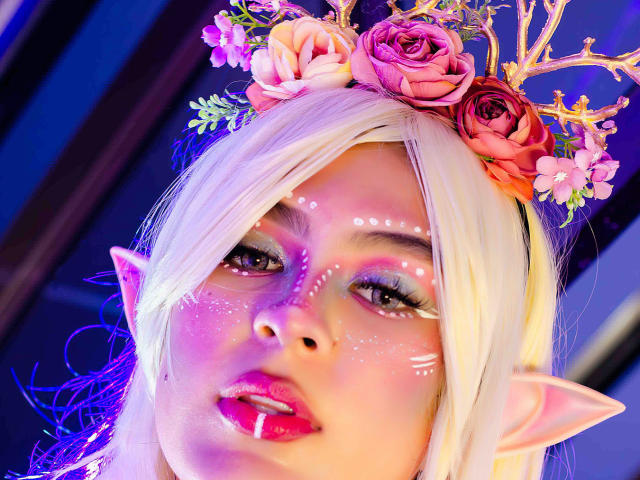 SamanthaEva69 with blonde hair, pink flower crown, and elf ears, has glitter on her face, pink lipstick, and glowing eyes, looking sexy with pointed ears and floral headpiece.