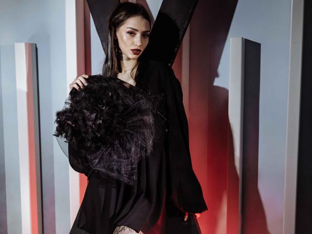SophieKitty with dark makeup, wearing a black dress and holding a feathery black accessory, standing in front of a V-shaped structure with red and gray lighting.