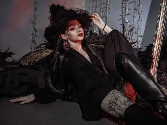 SophieKitty with black witch hat, dark lipstick, and long black hair, lying on black fabric, wearing black top, leather boots, and snake-patterned socks, looking seductive.
