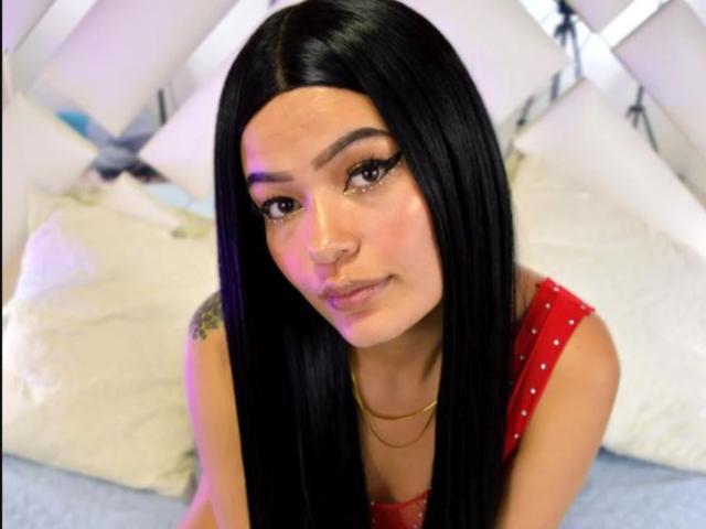 MelodyBellanova has long black hair, a nose ring, and a tattoo on her right shoulder. She's wearing a red, sleeveless top with a gold necklace. Her ass is unseen.