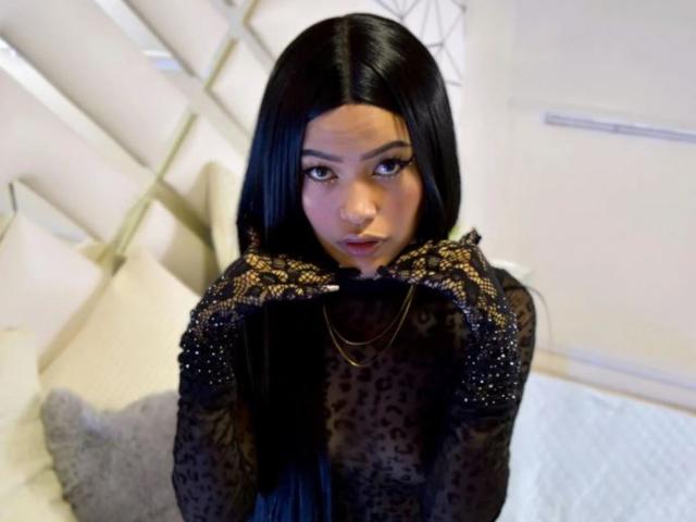 MelodyBellanova with long black hair, looking up with hands near face, wearing a black sheer top with gold embroidered gloves, and a gold necklace, in a bedroom.
