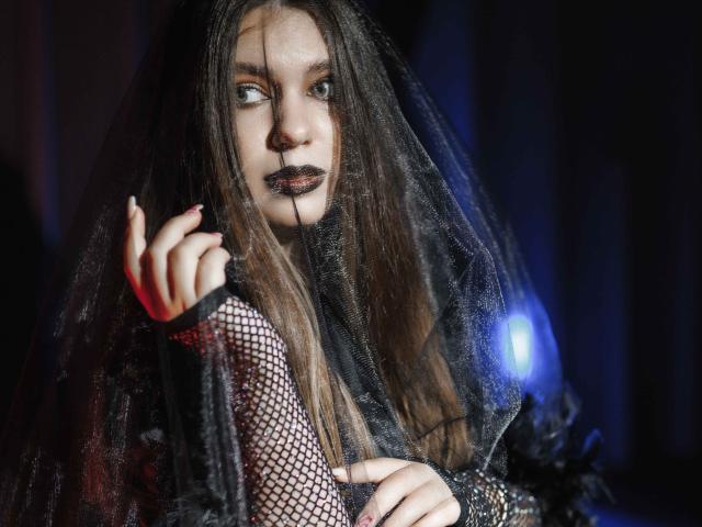 MilanaKitty with long brown hair, black mesh veil, fishnet sleeves, dark lipstick, and red nail polish, gazing intensely with a blue light in the background.