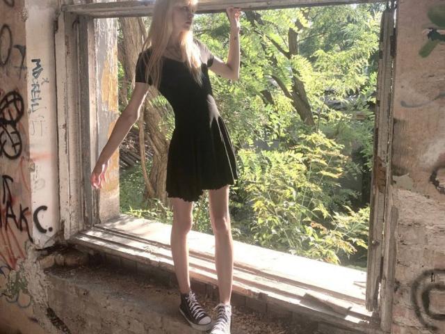 Lialit stands in a sunlit, graffiti-covered window, wearing a black dress and black-and-white sneakers, with long blonde hair and a cute, slender build.