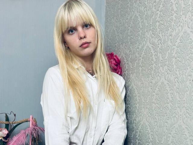 Lialit, met blond haar, blauwe ogen en een witte blouse, staat tegen een muur met een patroon, en kijkt iets opzij met een neutrale uitdrukking. In de achtergrond staan roze bloemen..