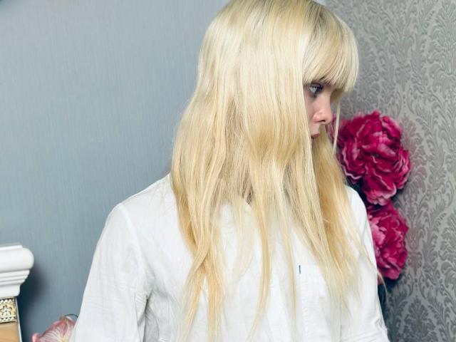 Lialit, con cabello rubio largo y liso, con flequillo, vestida con una camisa blanca, mirando hacia un lado con una expresión ligeramente triste, cerca de flores rosadas..