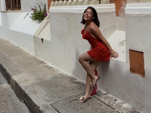 GiorginaR has dark hair, red dress, and red heels, leaning against a white wall with a smile, showing her ass and crossed legs, wearing open-toed sandals.